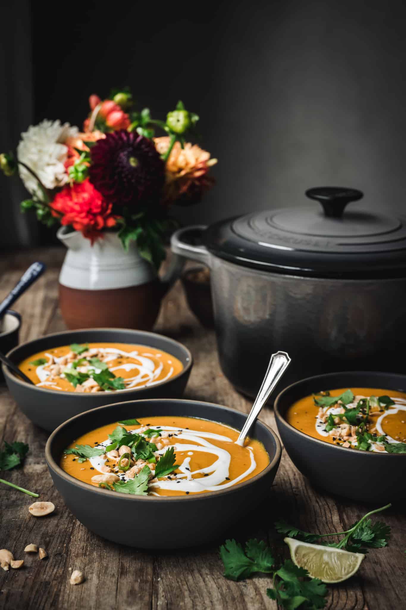 side view of three bowls of vegan coconut curry squash soup on wood table with fall flowers in background