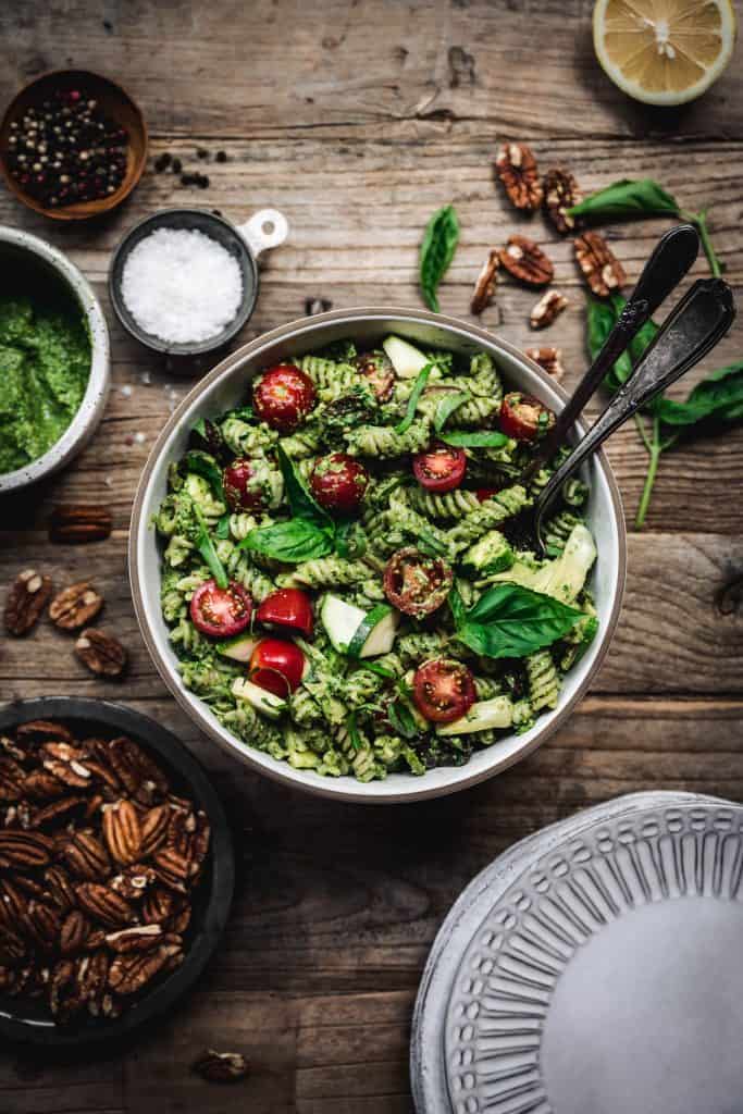 overhead view of pesto pasta salad with summer vegetables in white bowl with serving spoons