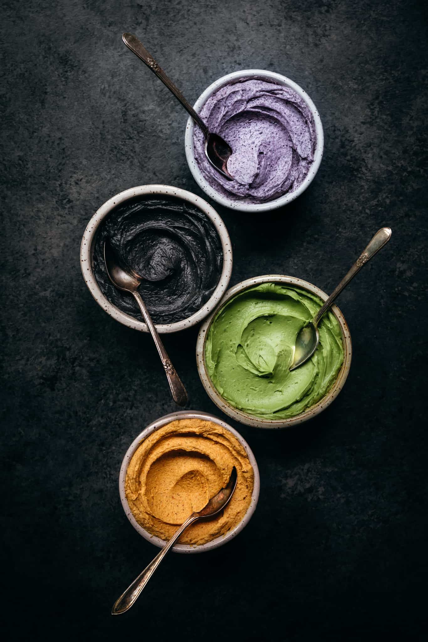 overhead of four bowls of frosting colored with natural food coloring from acai, beet, matcha and charcoal
