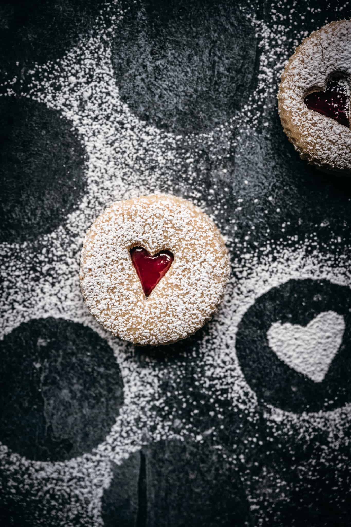 overhead view of linzer cookie with small heart cut out on black background with powdered sugar