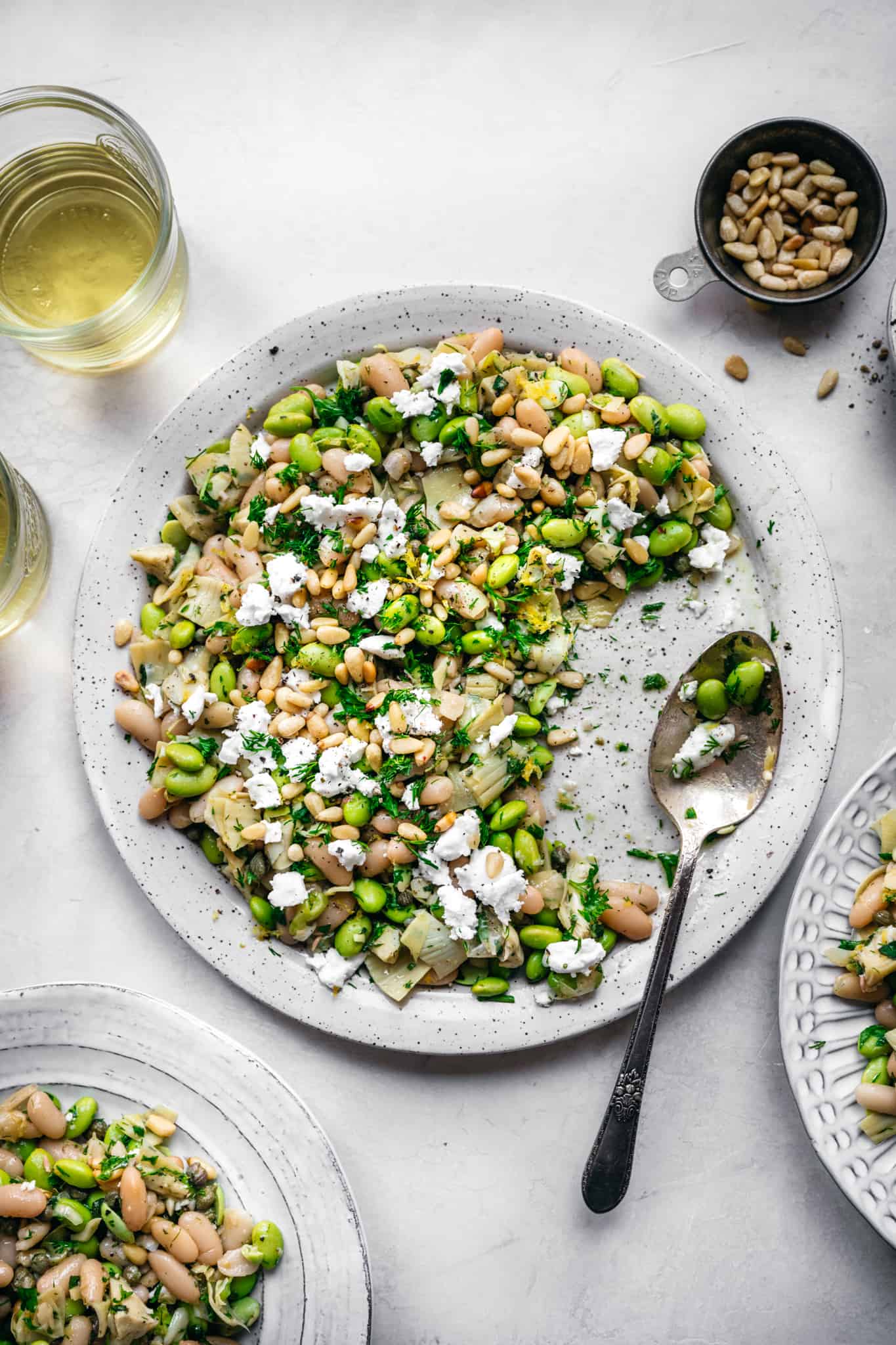 Overhead view of white bean salad with edamame, artichokes, pine nuts and feta on a white plate with serving spoon.