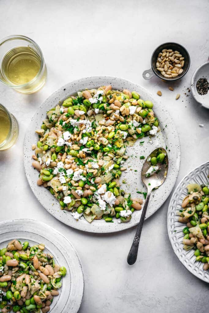overhead view of white bean salad with edamame, artichokes, pine nuts and feta on a white plate with serving spoon and white wine