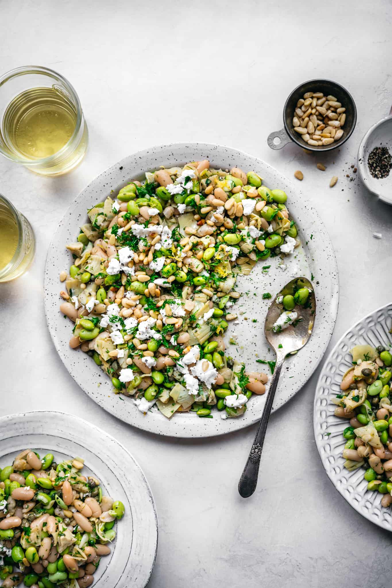 overhead view of white bean salad with edamame, artichokes, pine nuts and feta on a white plate with serving spoon and white wine