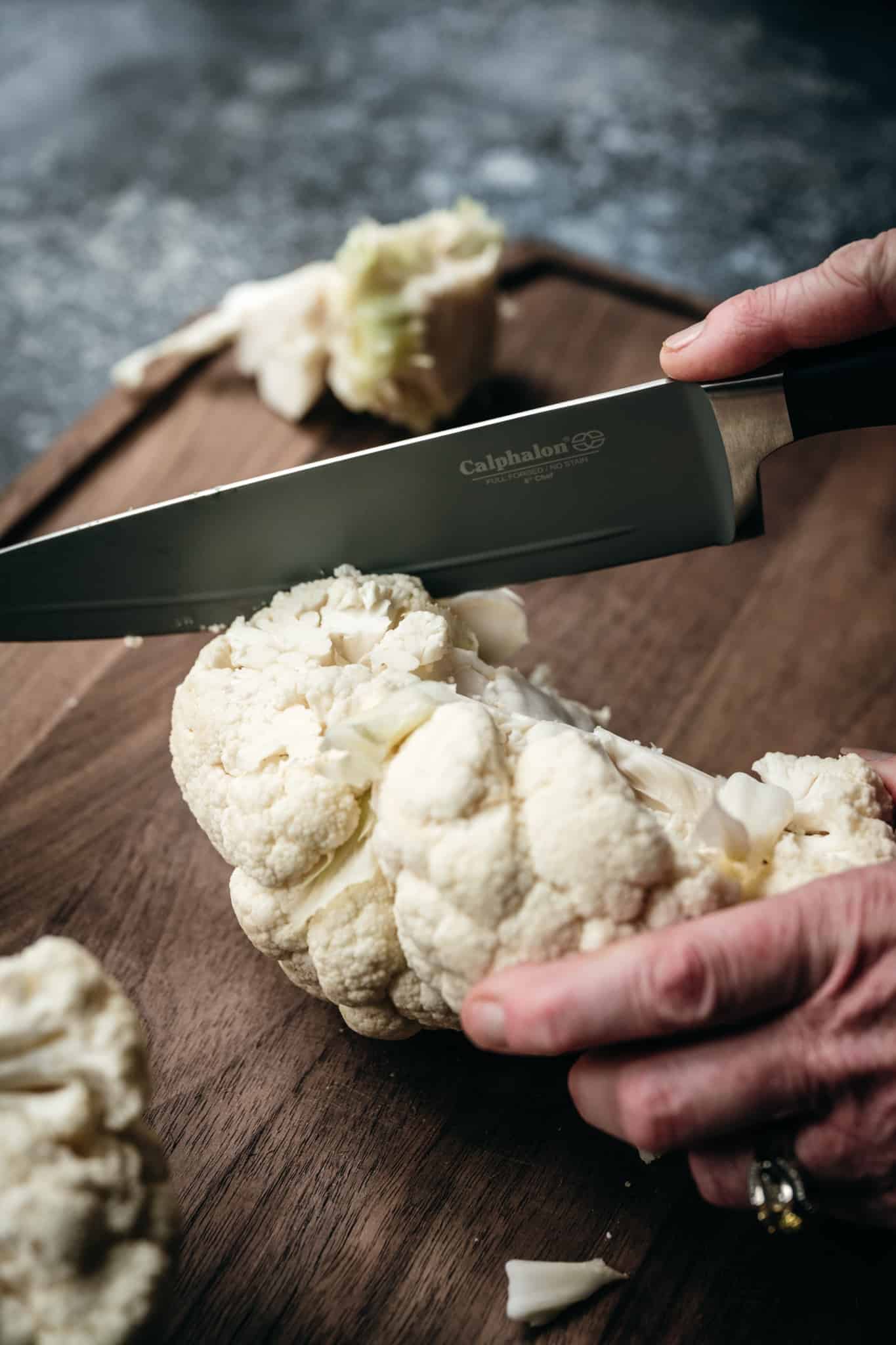 45 degree angle of a person slicing a head of cauliflower on a wooden cutting board with a chef's knife