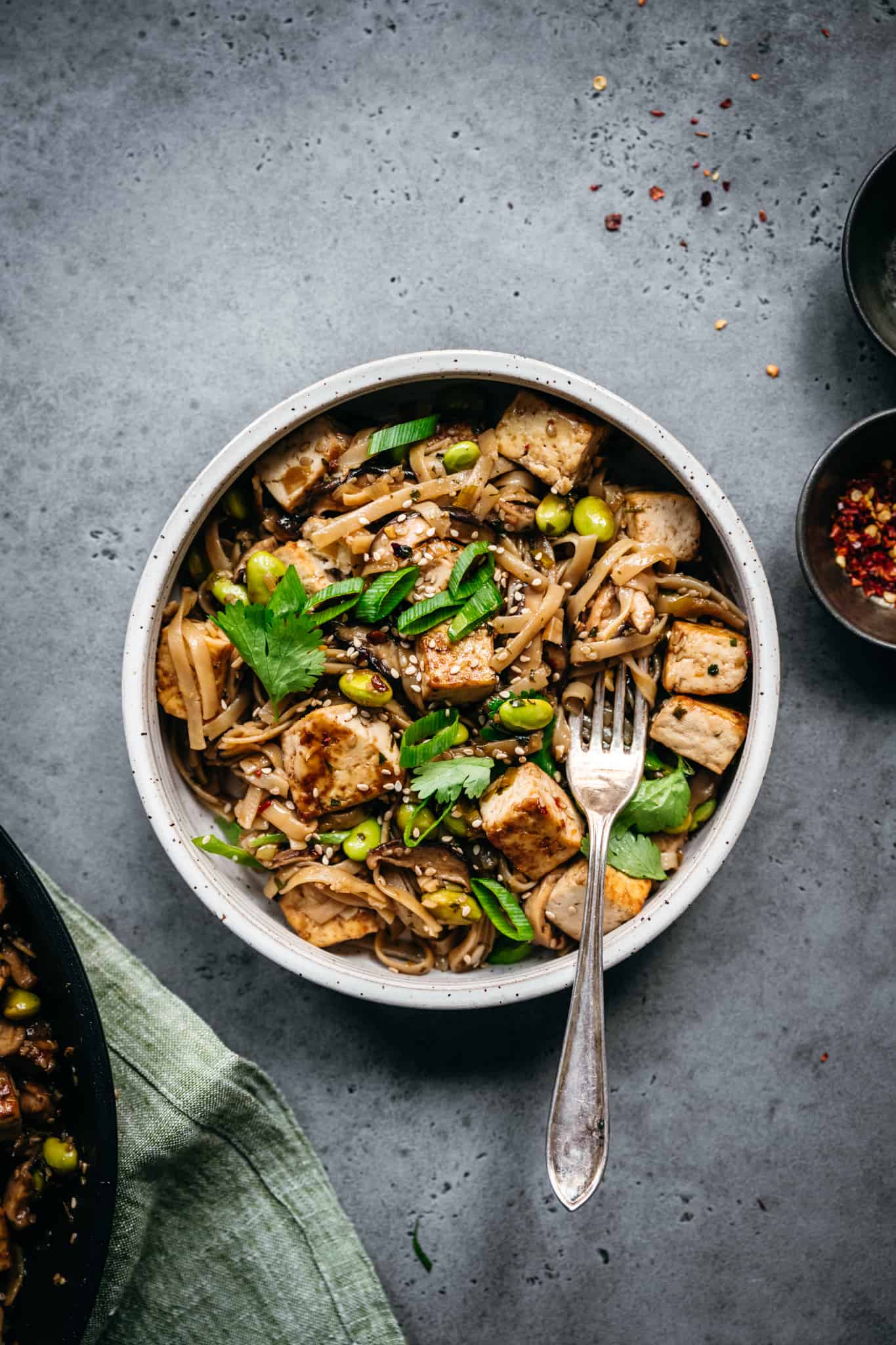 close up view of vegan rice noodles with crispy tofu, mushrooms and edamame in white bowl with fork
