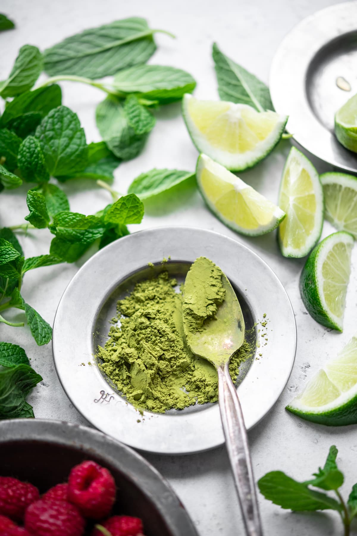 side view of matcha powder in small bowl next to mint, lime slices and raspberries