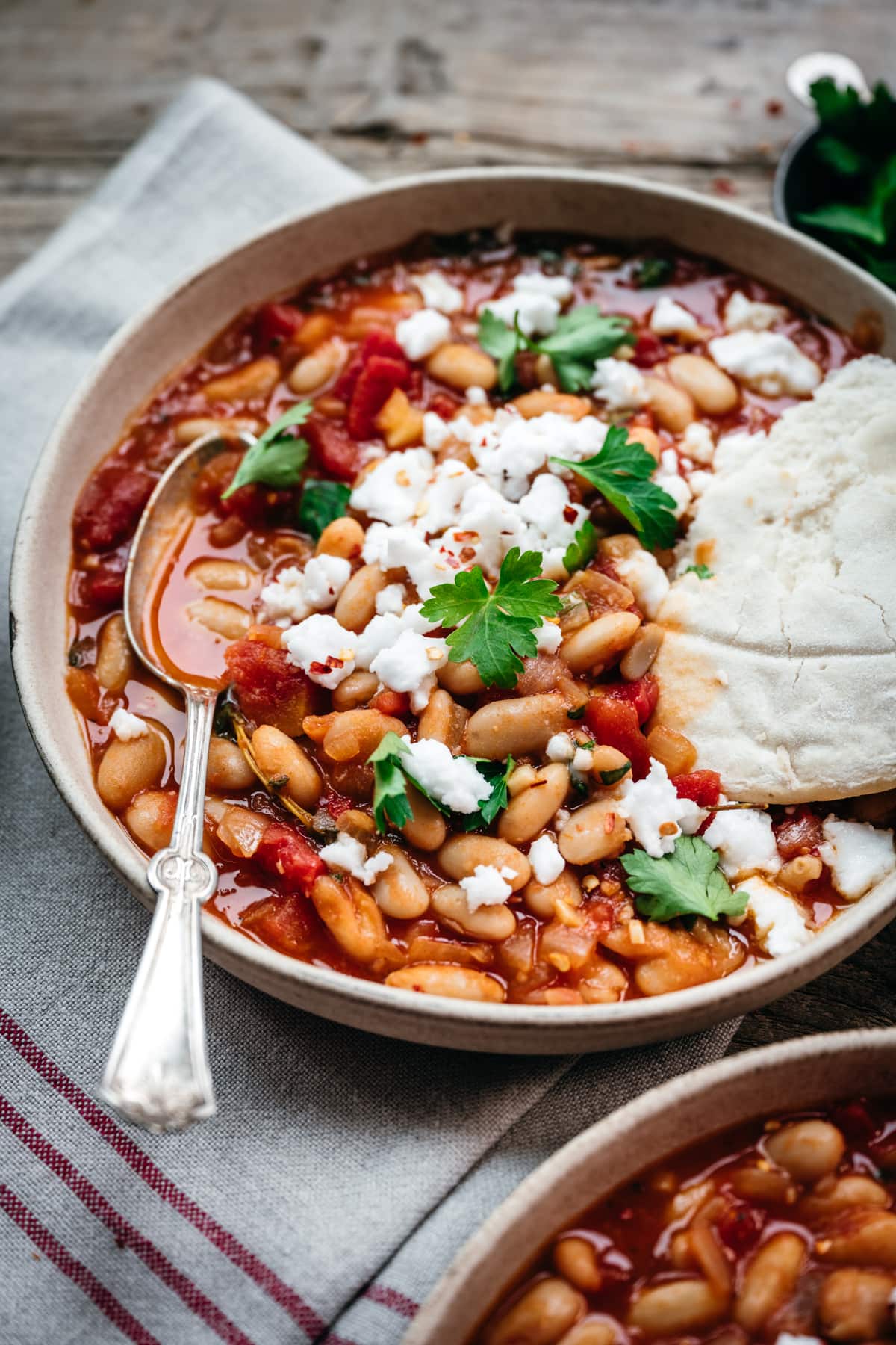 close up side view of tomato white bean stew in a bowl with feta and pita bread