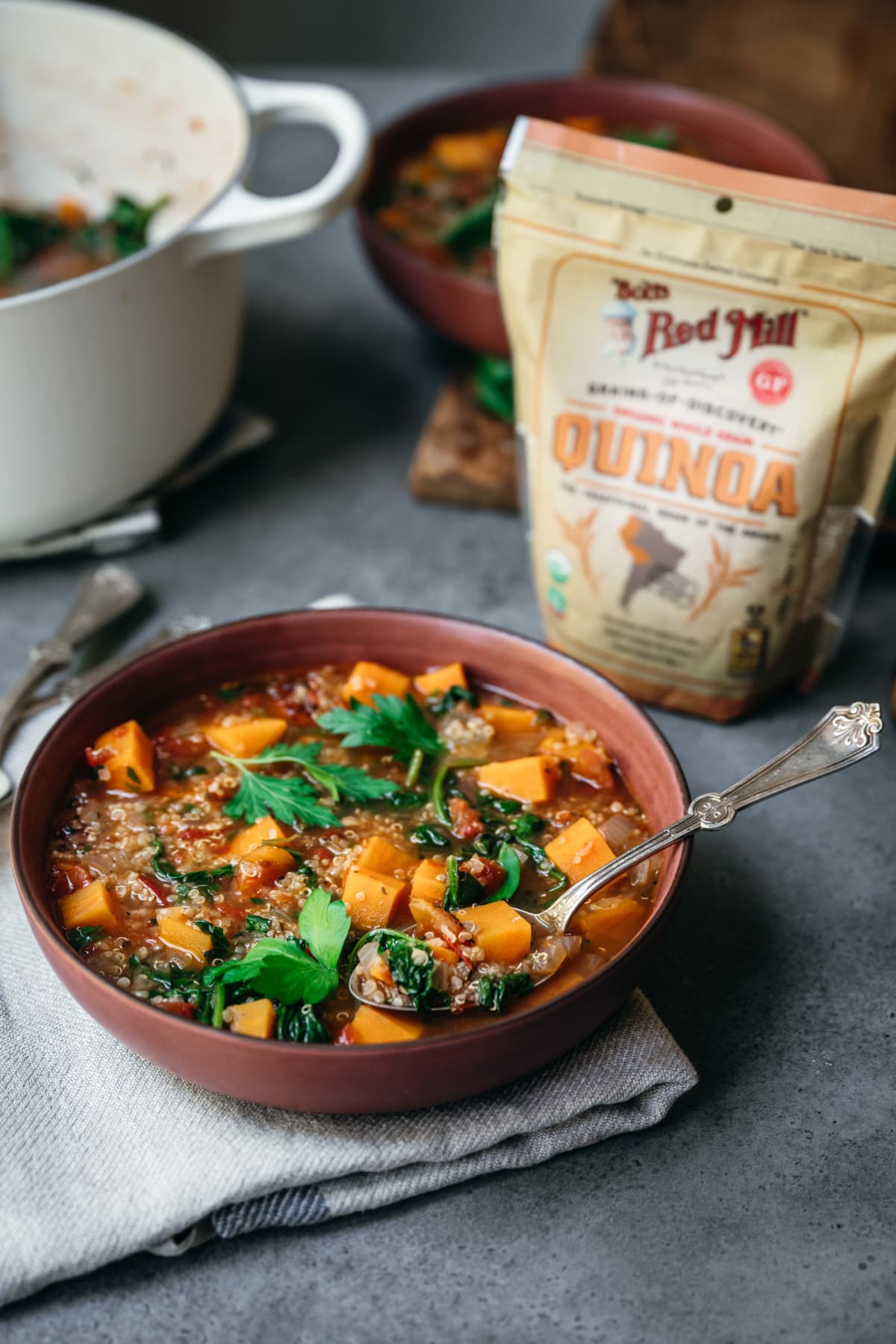 side view of vegetable quinoa soup in red bowl with bob's red mill quinoa bag. 