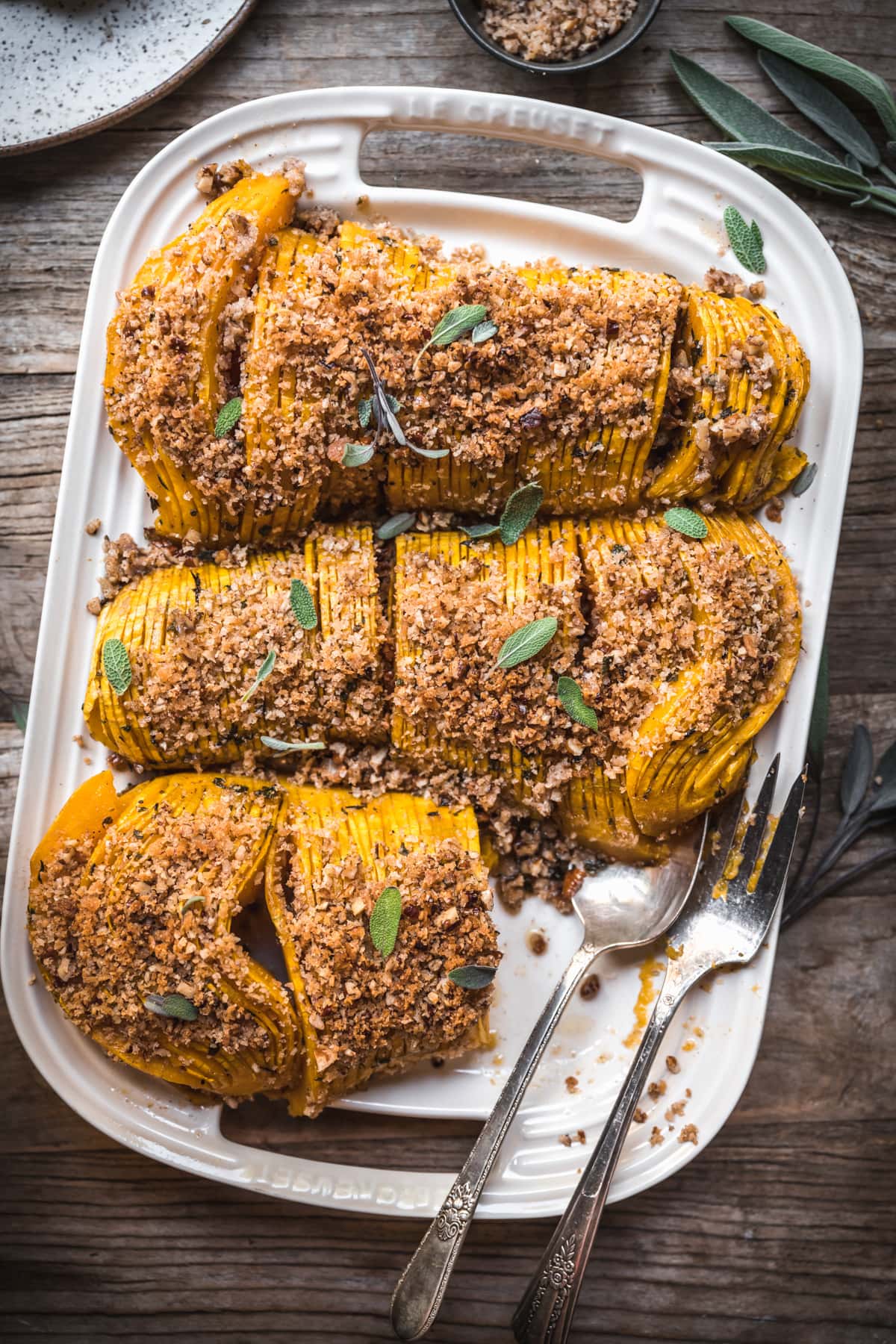 overhead view of hasselback butternut squash with breadcrumbs on a white platter with serving utensils.