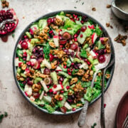 overhead view of vegan waldorf salad with walnut brittle and pomegranate in a bowl with serving utensils.