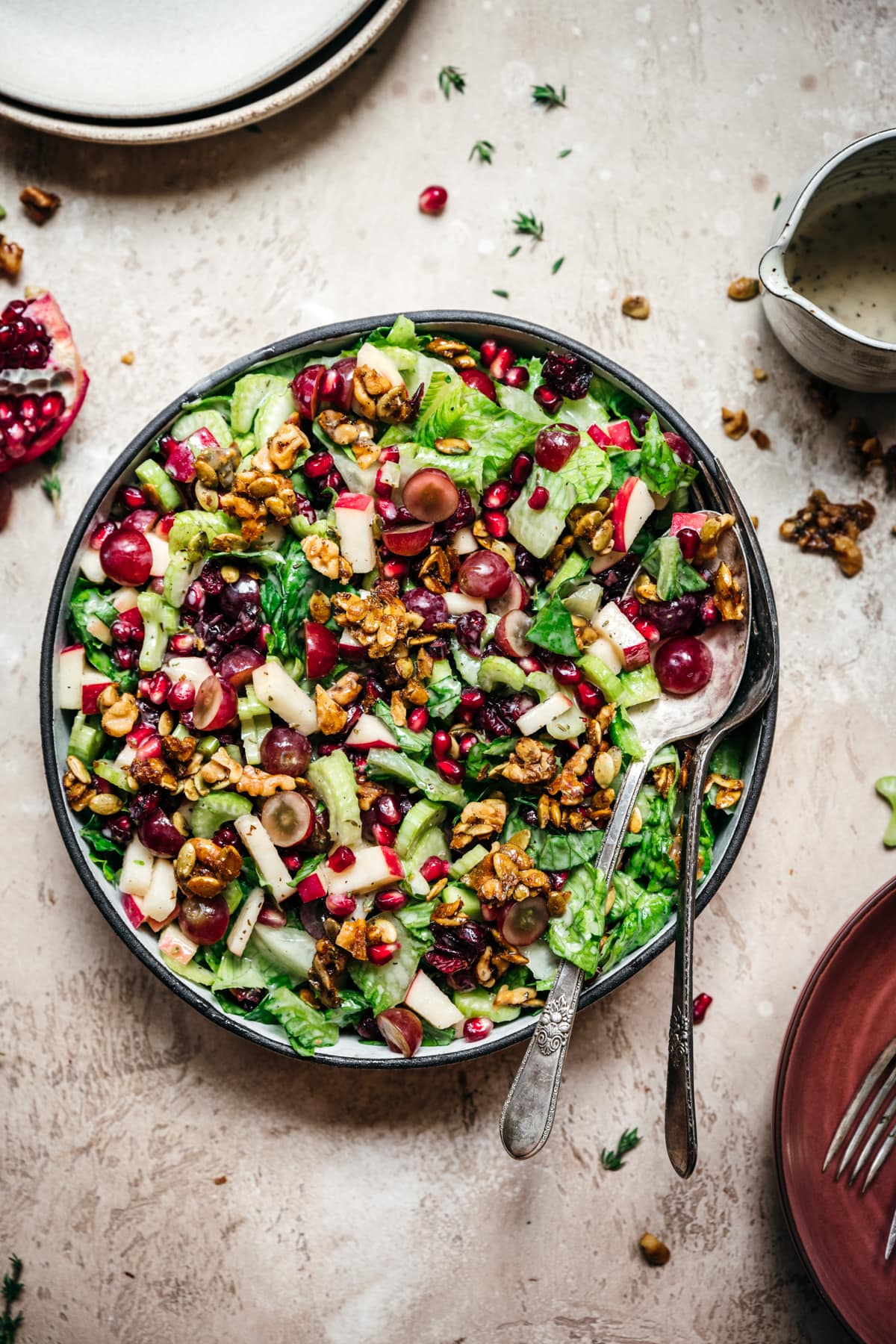 overhead view of vegan waldorf salad with walnut brittle and pomegranate in a bowl with serving utensils.