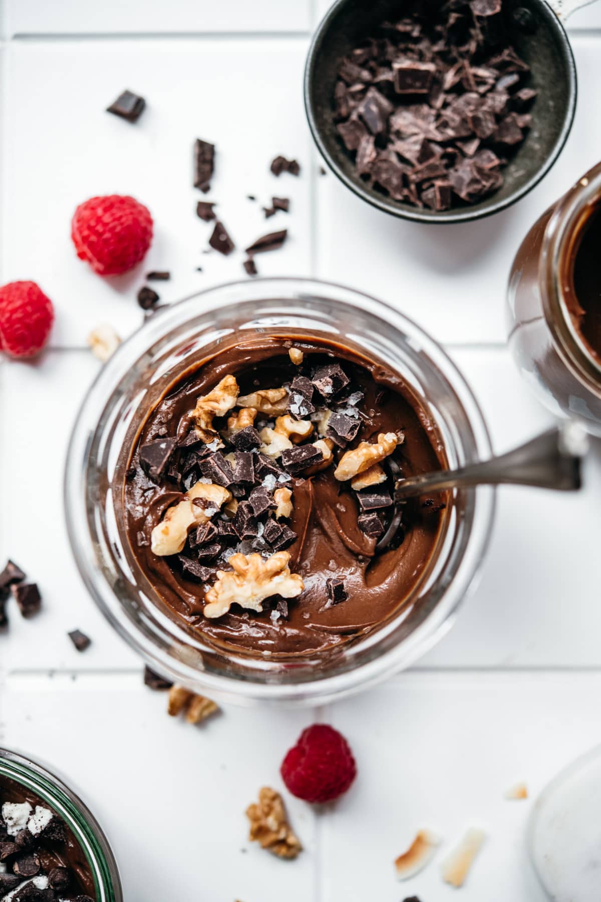 overhead view of jar of vegan avocado chocolate mousse with nuts on white tile. 