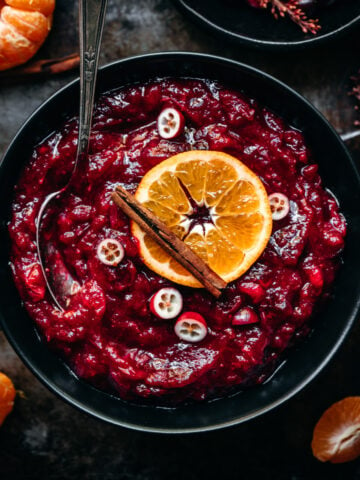 overhead view of cranberry sauce in a black bowl garnished with orange slice and cinnamon stick.