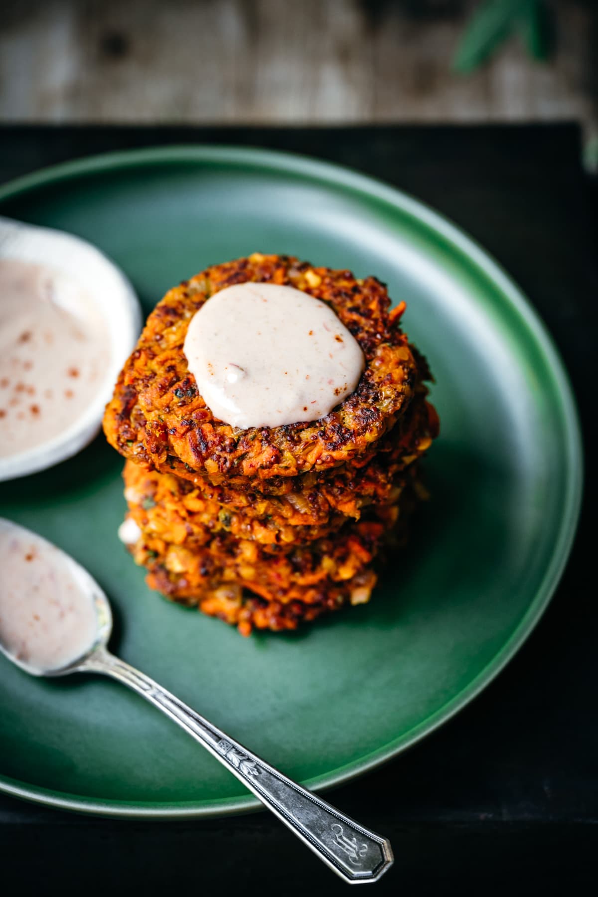 side view of a stack of carrot fritters on a green plate with yogurt sauce on top.