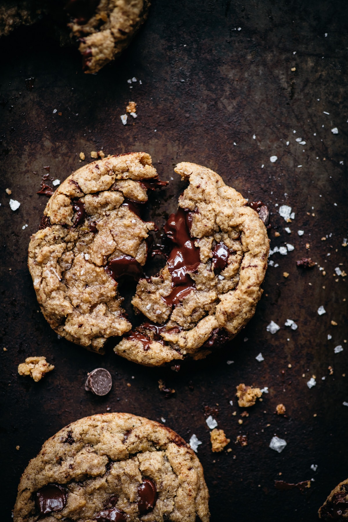 close up overhead view of melty vegan chocolate chip cookie broken in half on rustic sheet pan. 