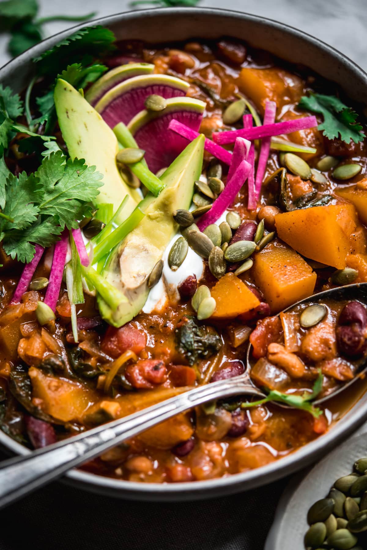 overhead view of bowl of vegan pumpkin chili topped with avocado slices, sour cream, pumpkin seeds and watermelon radish.