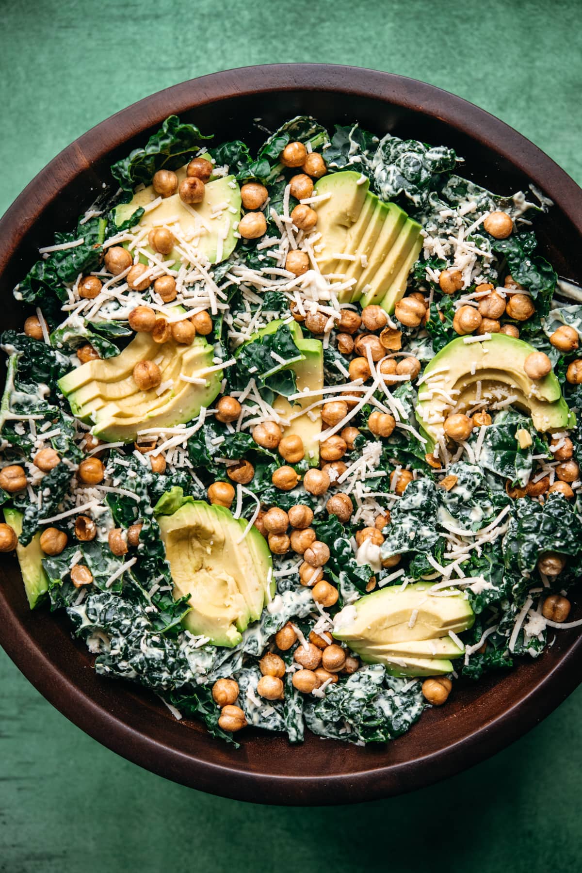 overhead view of vegan kale caesar salad in a large wood bowl topped with avocado and crispy chickpeas. 