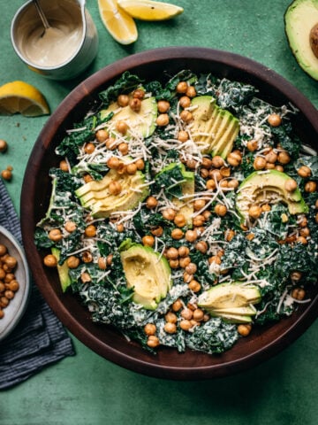 overhead view of vegan kale caesar salad in a large wood bowl topped with avocado and crispy chickpeas.