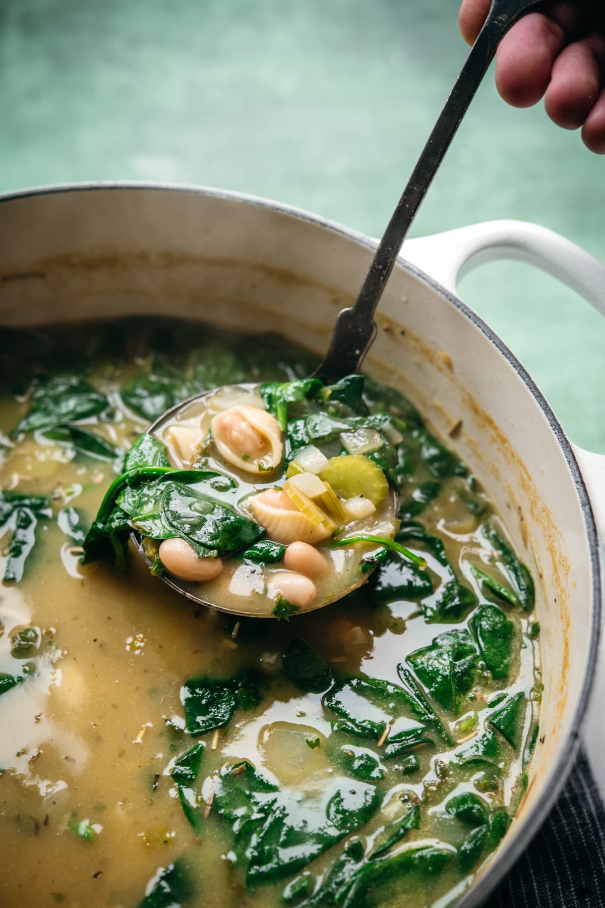 close up view of person holding ladle of white bean spinach soup above soup pot. 