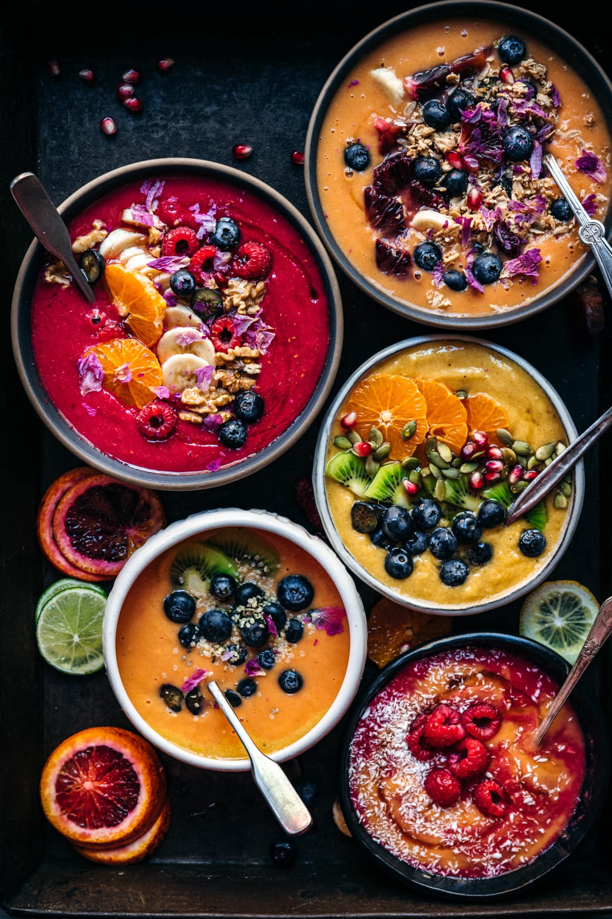 overhead view of multiple red, orange and yellow smoothie bowls with fresh fruit and granola toppings. 