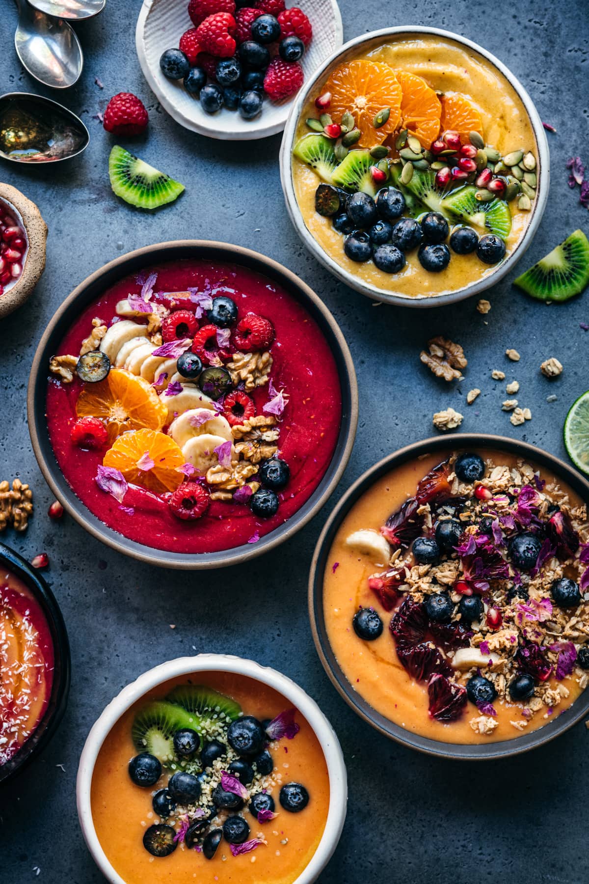 overhead view of multiple red, orange and yellow smoothie bowls with fresh fruit and granola toppings. 