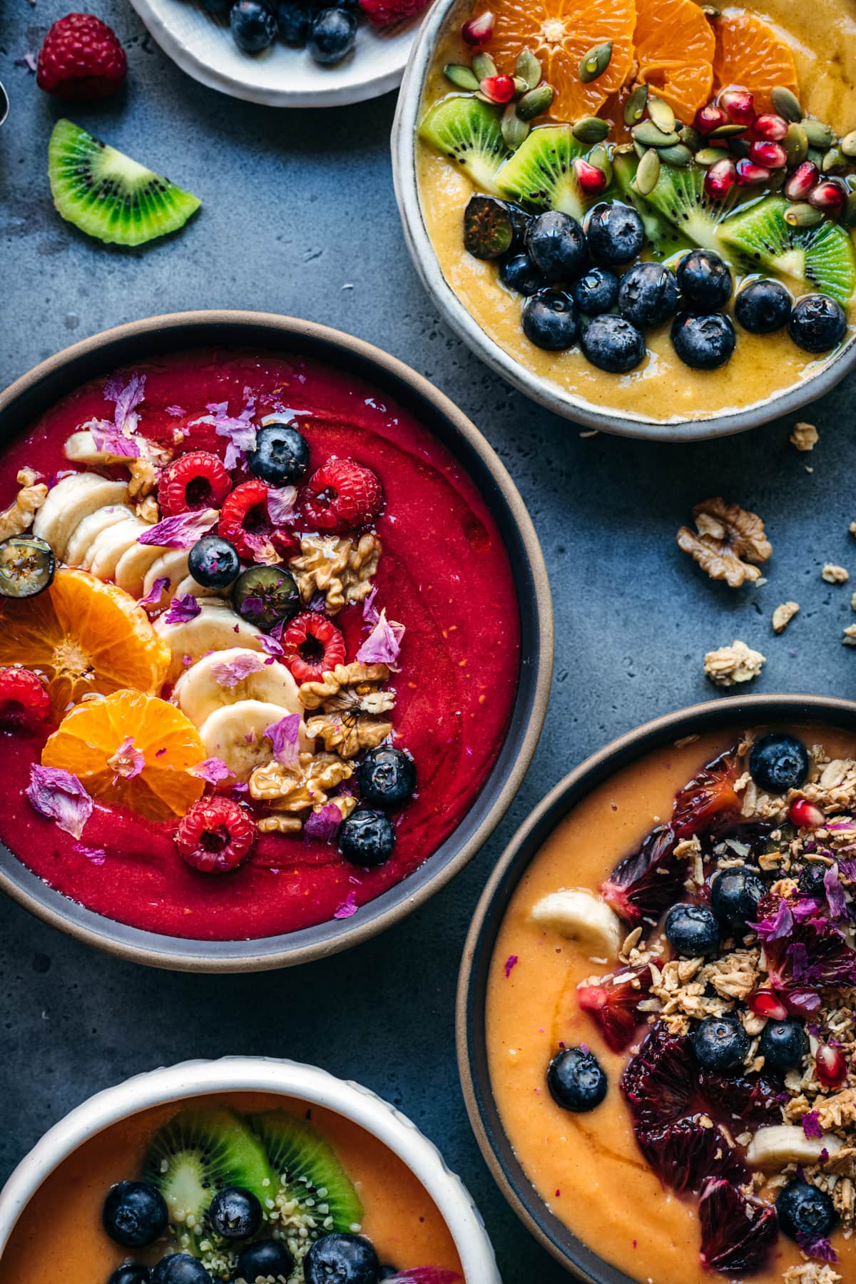 overhead view of multiple red, orange and yellow smoothie bowls with fresh fruit and granola toppings.