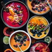 overhead view of multiple red, orange and yellow smoothie bowls with fresh fruit and granola toppings.