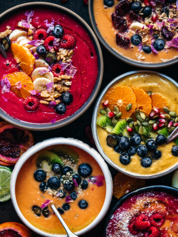 overhead view of multiple red, orange and yellow smoothie bowls with fresh fruit and granola toppings.