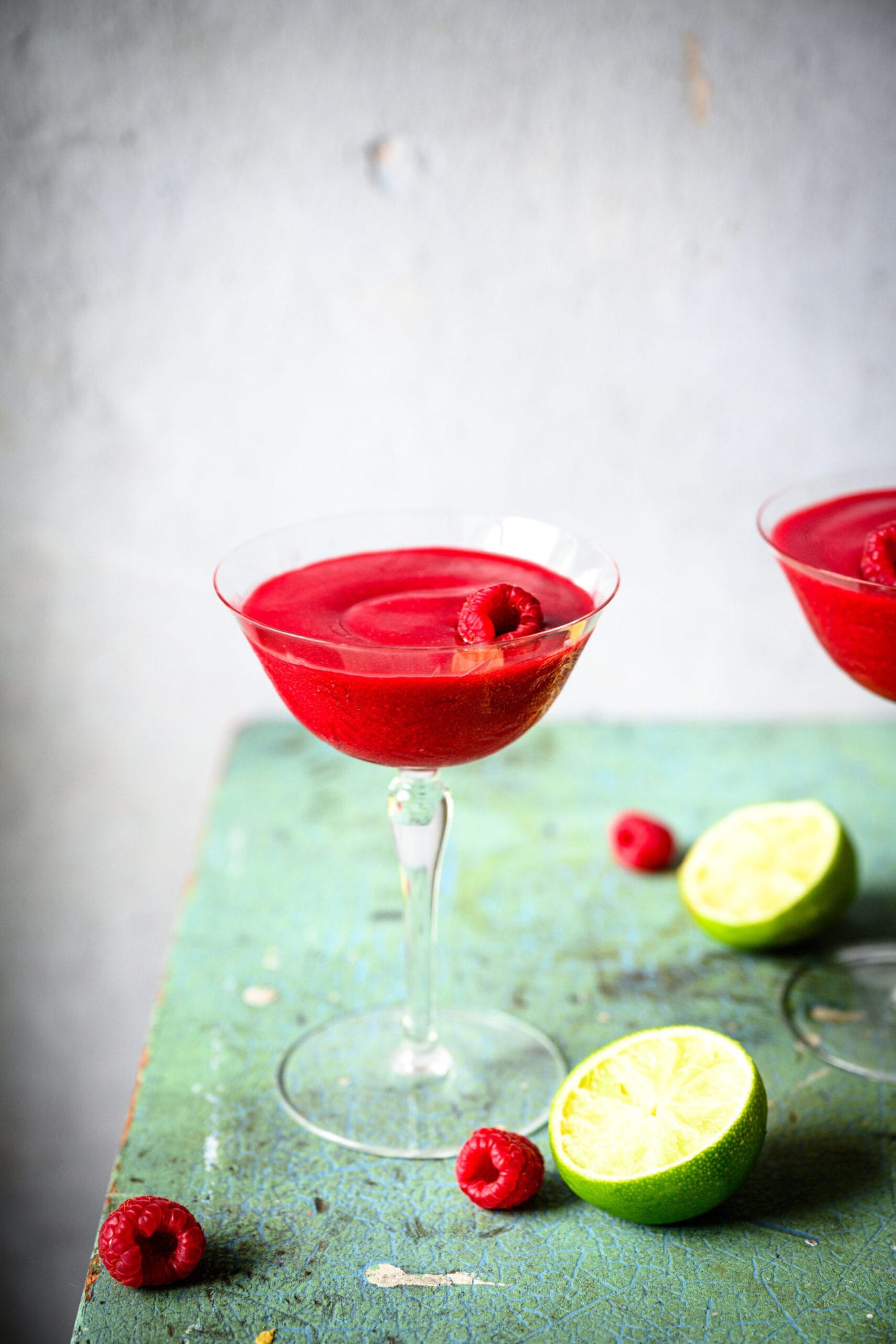 side view of frozen raspberry daiquiri in coupe glass with fresh raspberries and lime slices. 