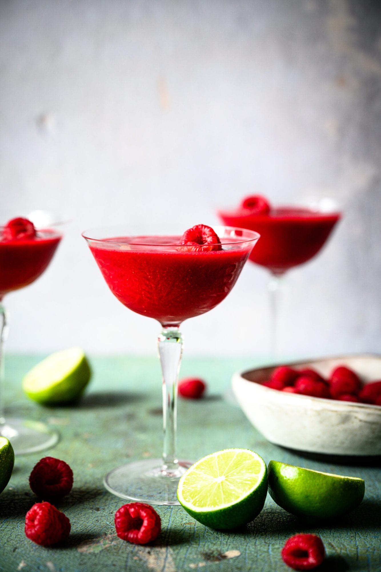 side view of frozen raspberry daiquiri in coupe glass with fresh raspberries and lime slices.