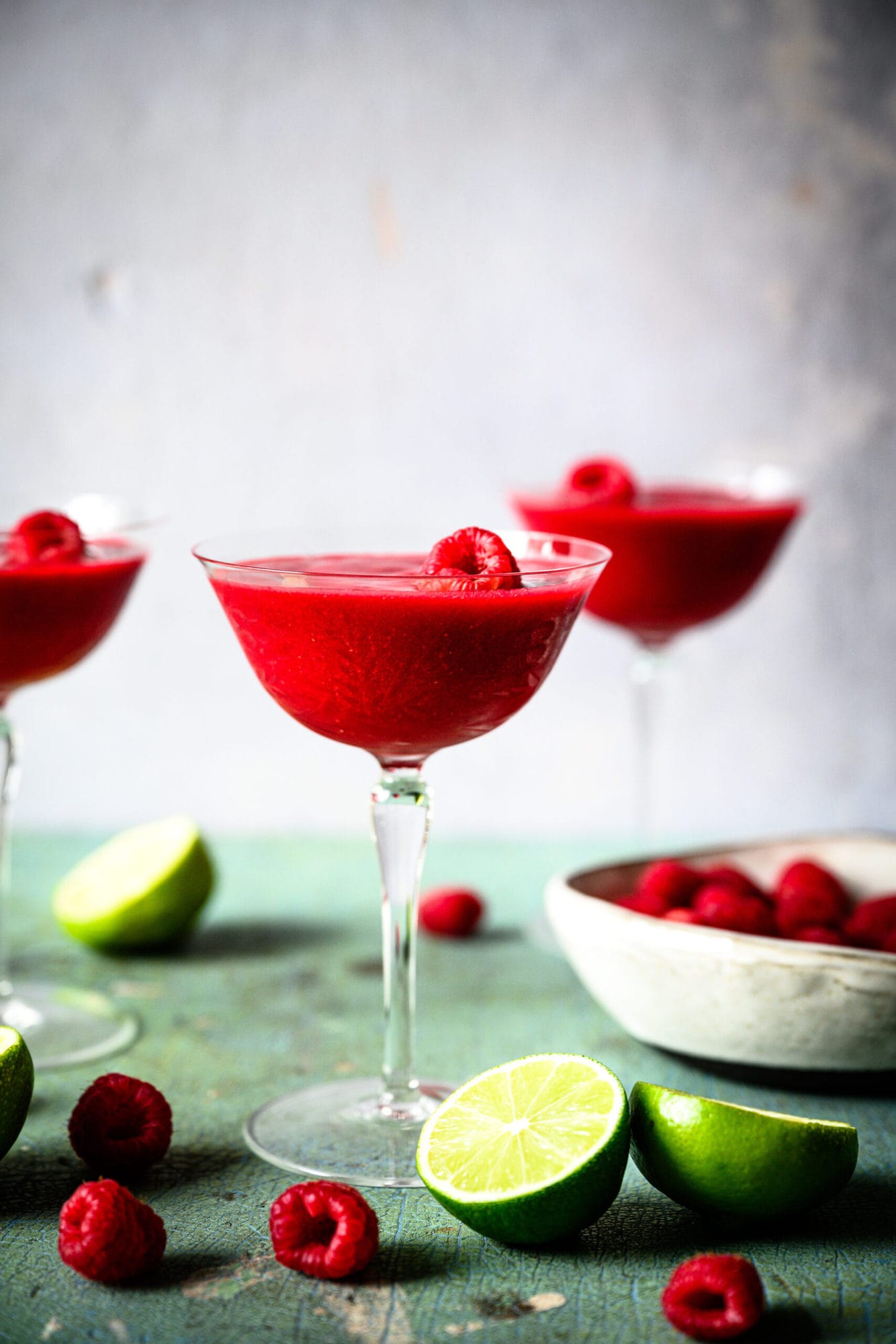 side view of frozen raspberry daiquiri in coupe glass with fresh raspberries and lime slices. 