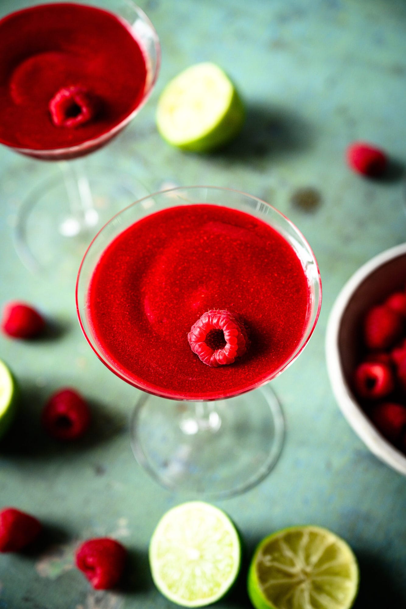 overhead view of frozen raspberry daiquiri in coupe glass with fresh raspberries and lime slices. 
