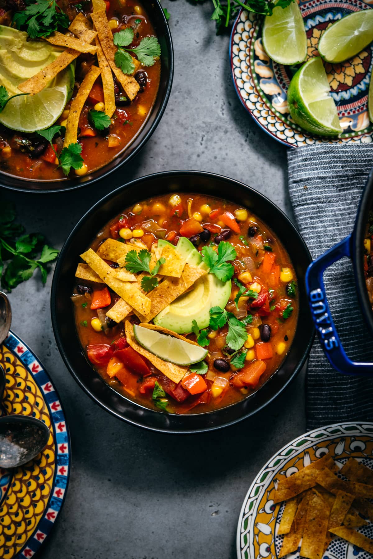 overhead view of vegan tortilla soup in black bowls topped with avocado, tortilla strips and lime. 