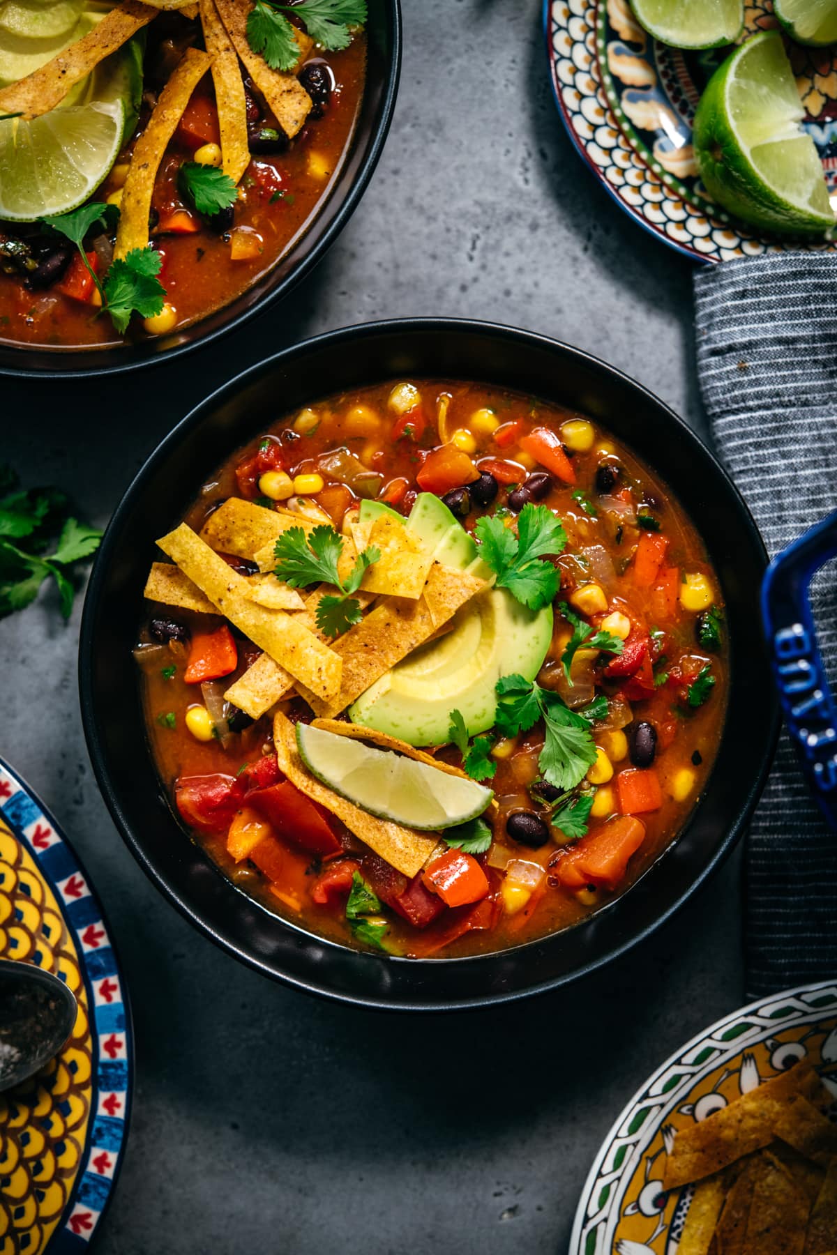 overhead view of vegan tortilla soup in black bowls topped with avocado, tortilla strips and lime. 