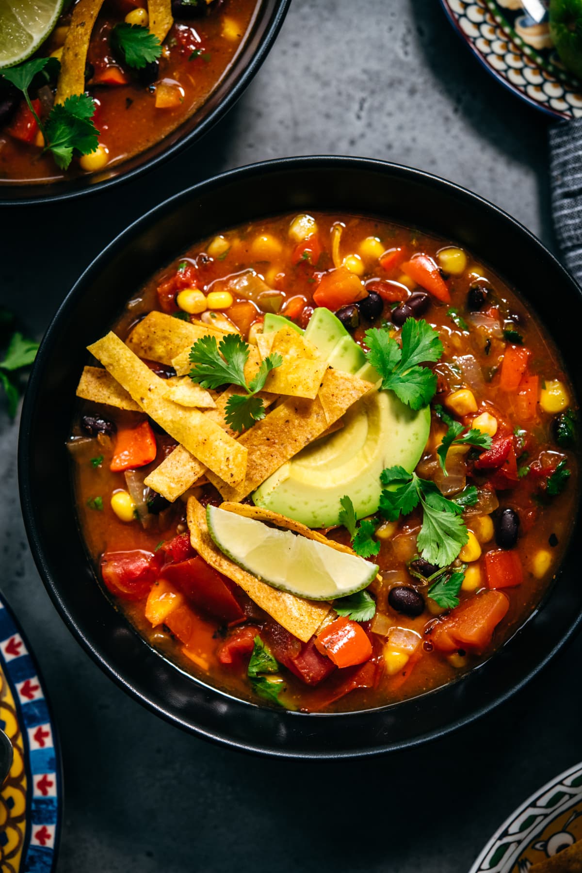 overhead view of vegan tortilla soup in black bowls topped with avocado, tortilla strips and lime. 