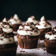 close up side view of vegan chocolate cupcake topped with vanilla frosting and chocolate shavings.