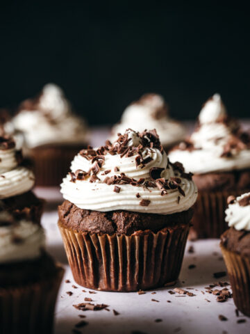 close up side view of vegan chocolate cupcake topped with vanilla frosting and chocolate shavings.