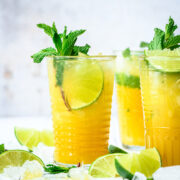 side view of mango mojitos in tall cocktail glasses garnished with mint and lime on white tile.