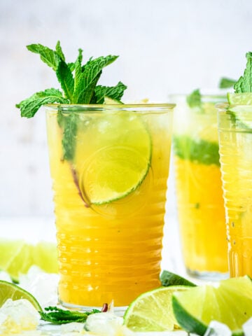 side view of mango mojitos in tall cocktail glasses garnished with mint and lime on white tile.