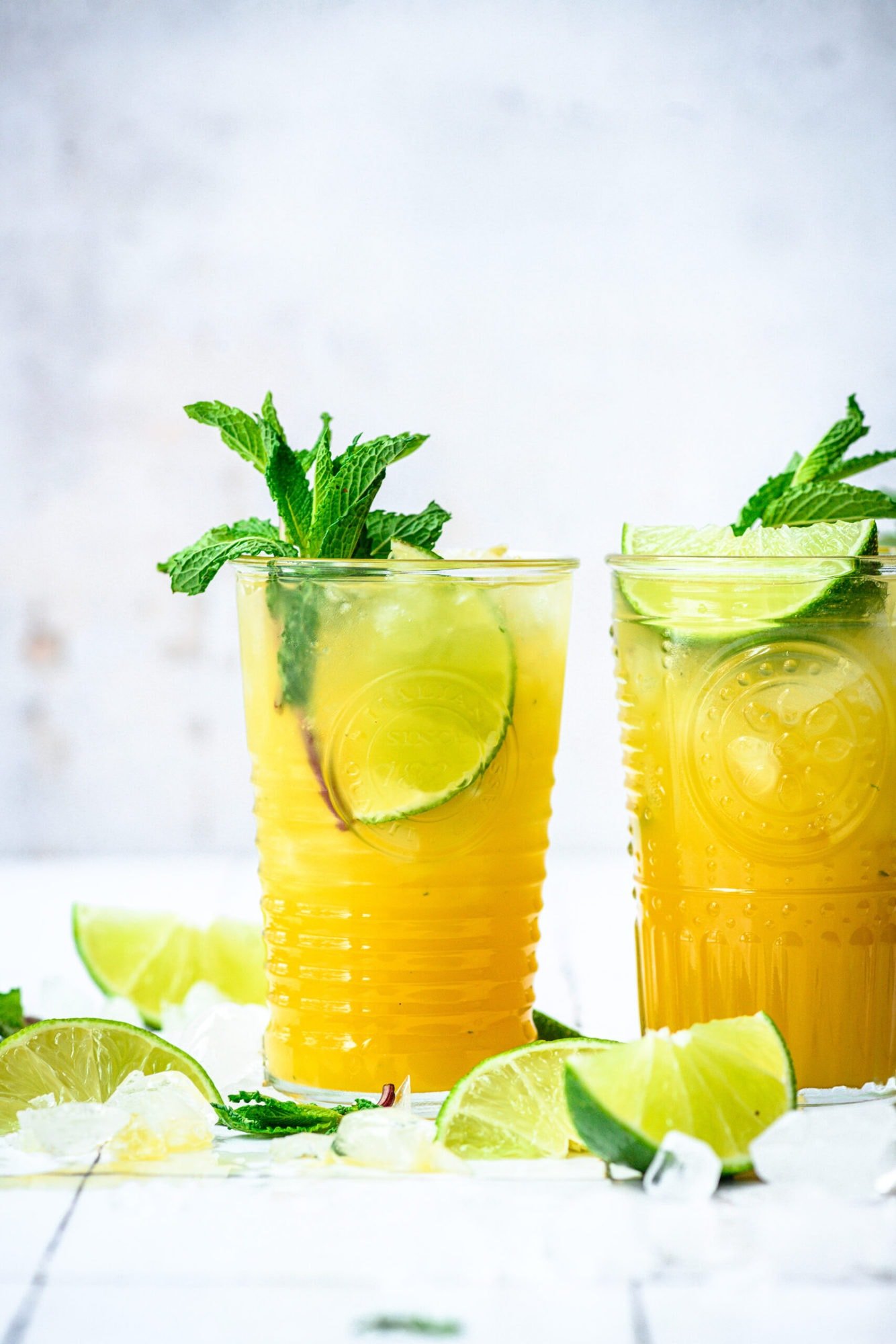 side view of mango mojitos in tall cocktail glasses garnished with mint and lime on white tile.