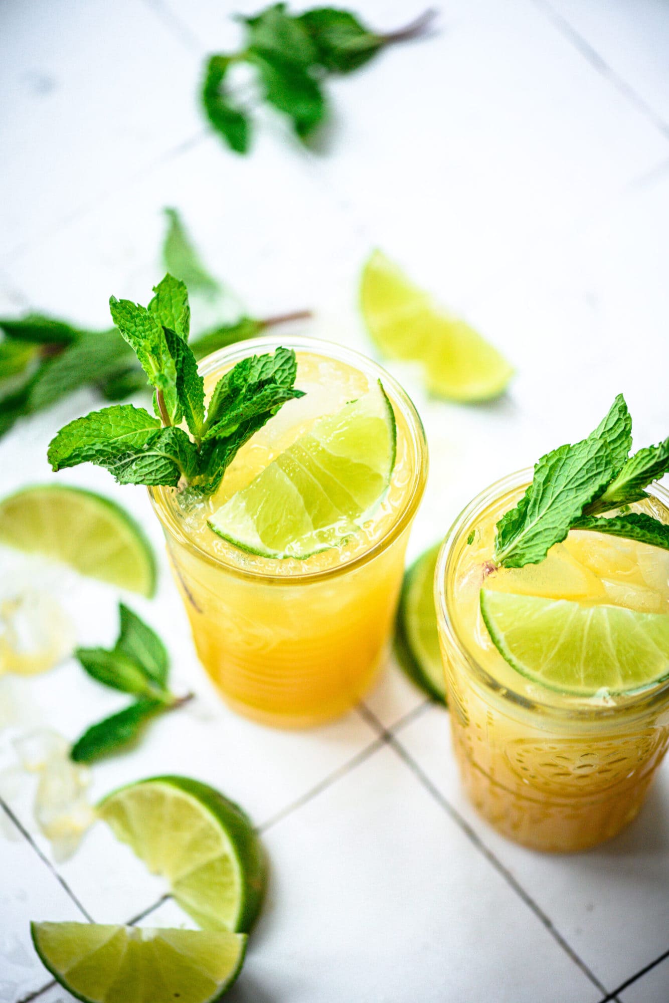 side view of mango mojitos in tall cocktail glasses garnished with mint and lime on white tile.