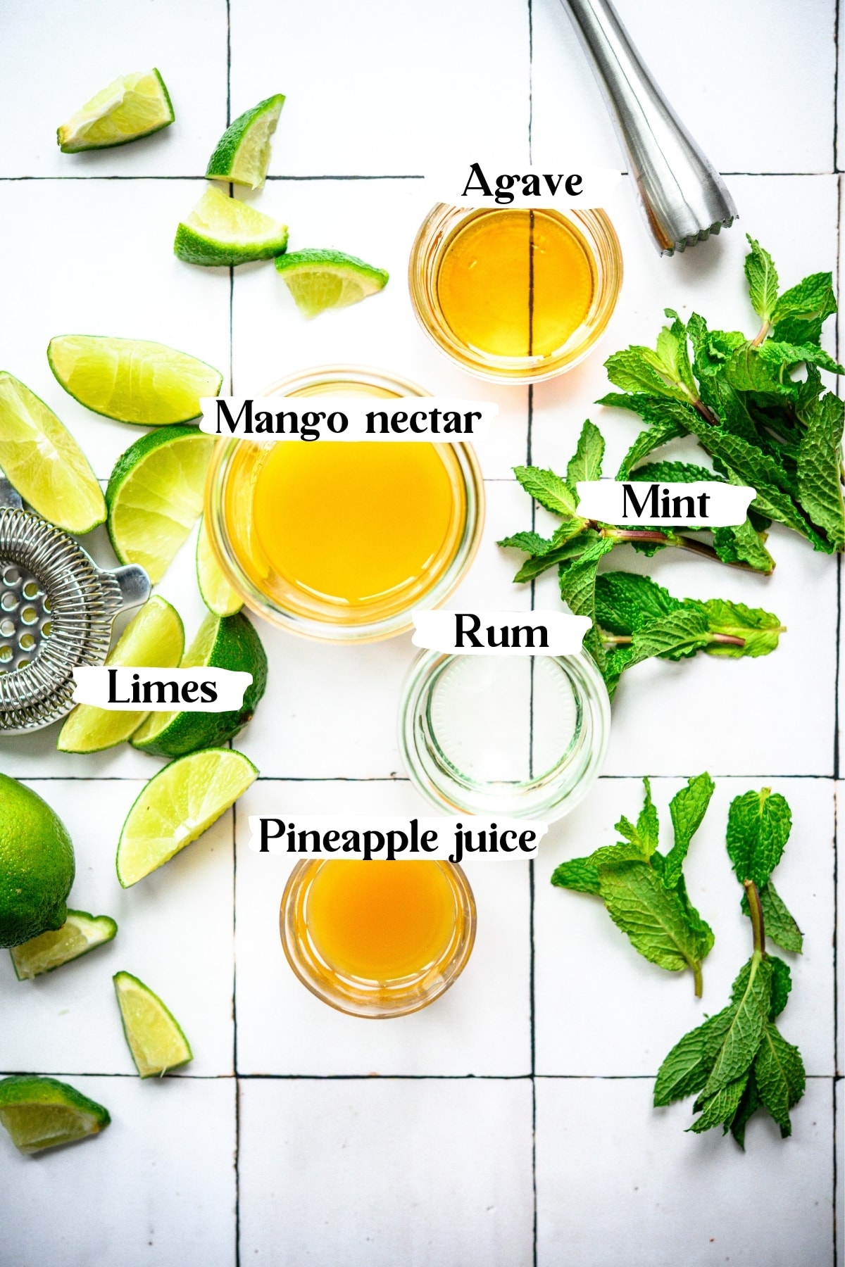 overhead view of mango nectar, rum, limes, mint, pineapple juice and agave in small bowls on white tile.