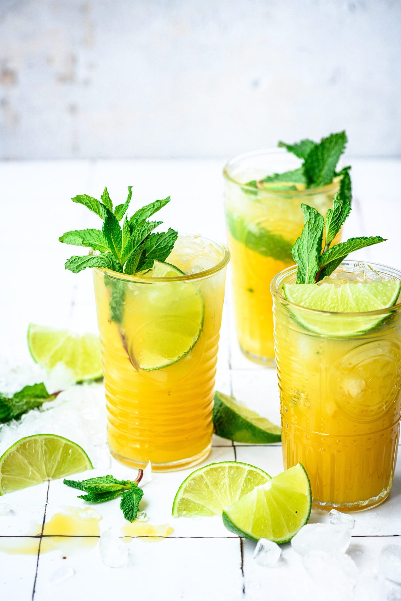 side view of mango mojitos in tall cocktail glasses garnished with mint and lime on white tile.