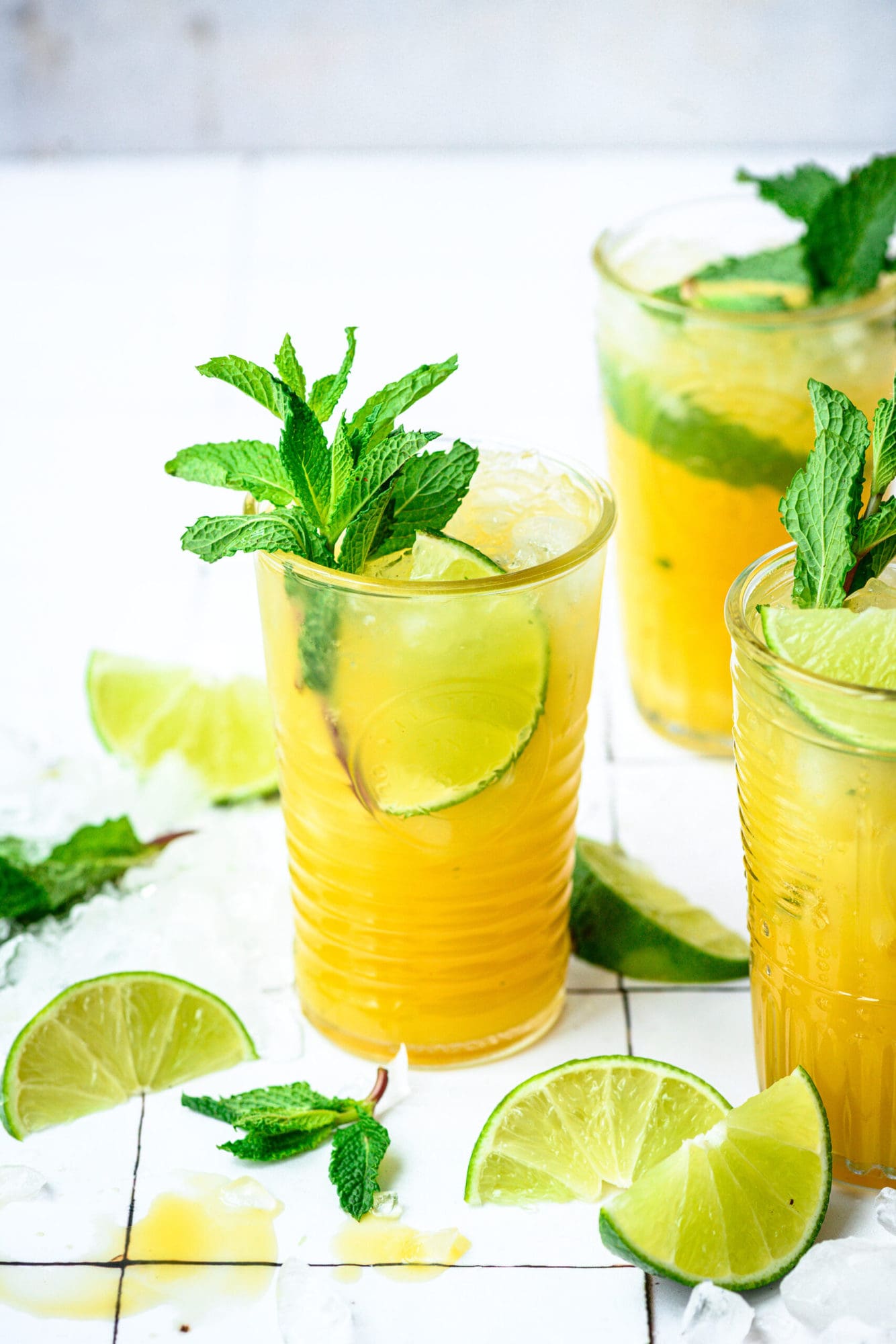 side view of mango mojitos in tall cocktail glasses garnished with mint and lime on white tile.