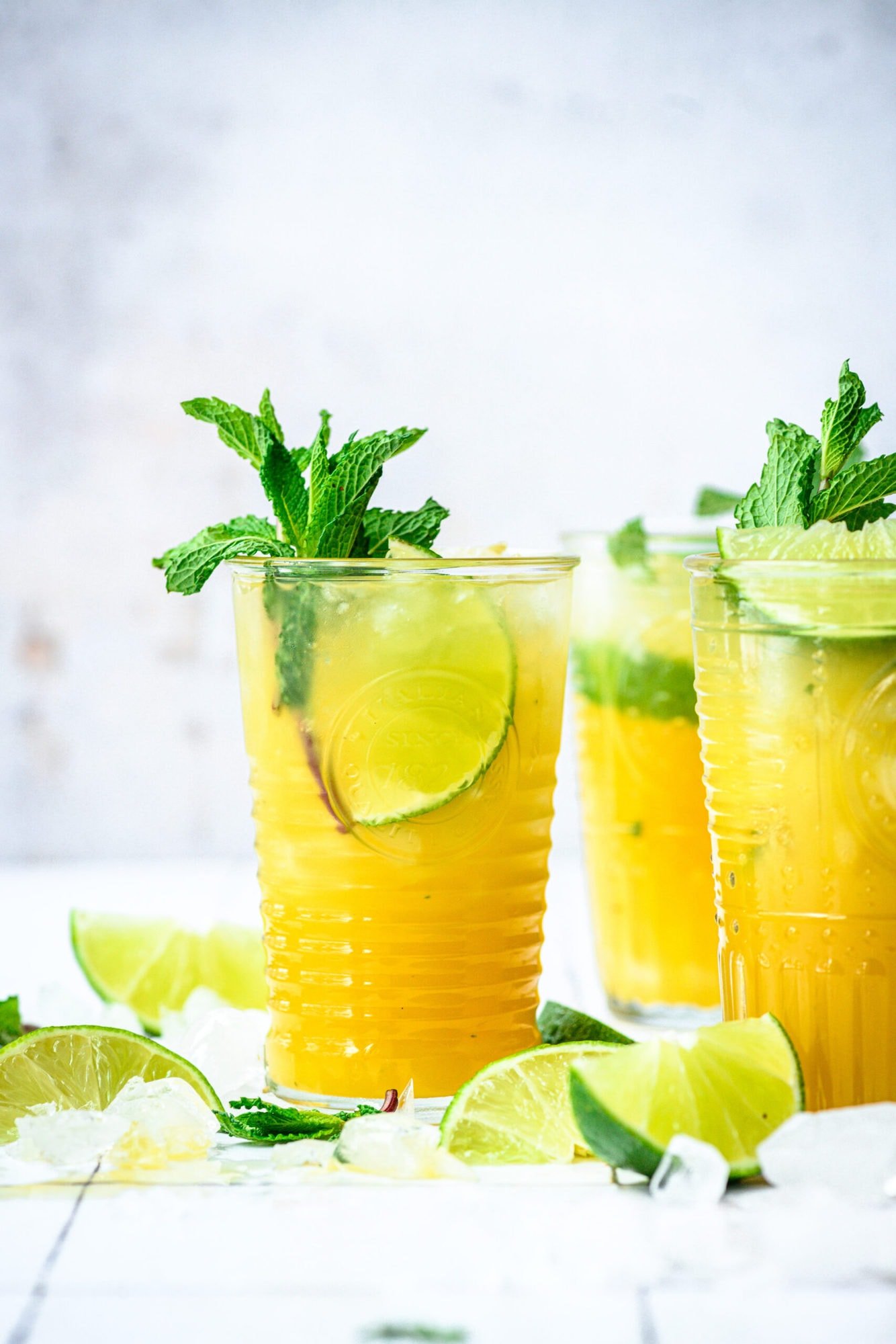 side view of mango mojitos in tall cocktail glasses garnished with mint and lime on white tile.