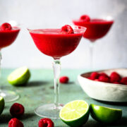 side view of frozen raspberry daiquiri in coupe glass with fresh raspberries and lime slices.