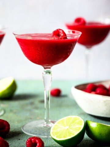 side view of frozen raspberry daiquiri in coupe glass with fresh raspberries and lime slices.