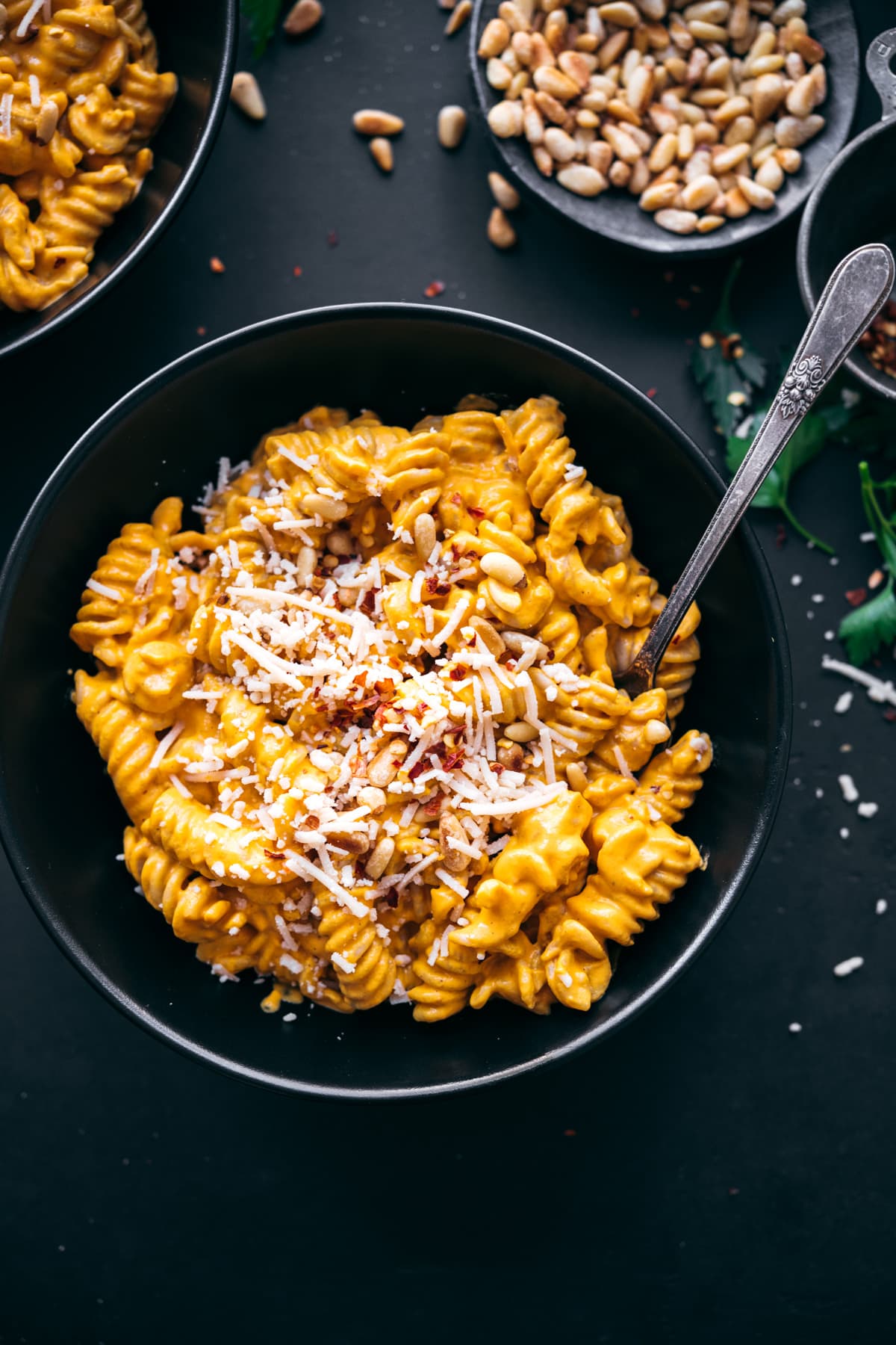 overhead view of creamy vegan roasted red pepper pasta in black bowl topped with parmesan, pine nuts and red pepper flakes. 