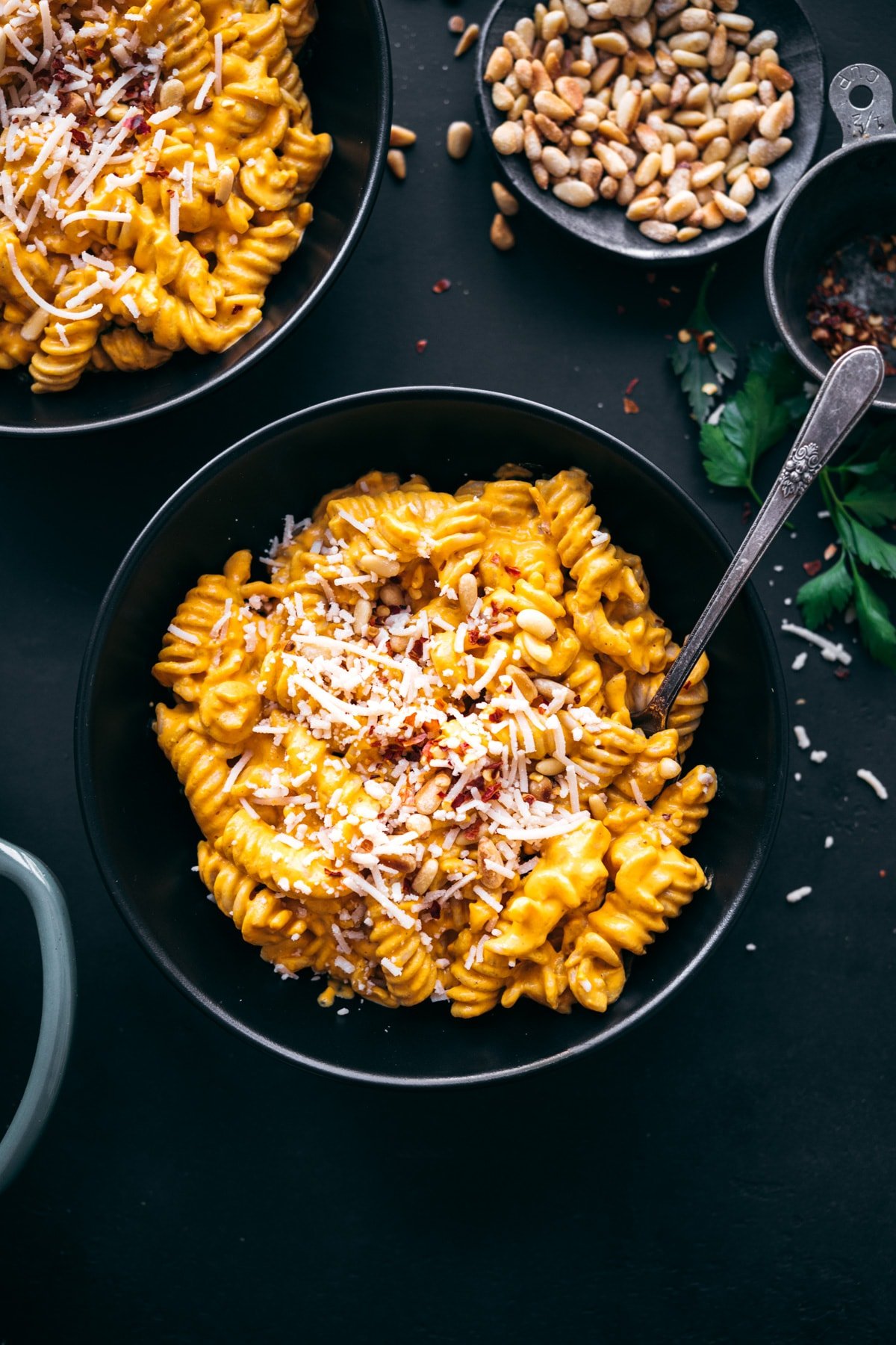overhead view of creamy vegan roasted red pepper pasta in black bowls topped with parmesan, pine nuts and red pepper flakes.