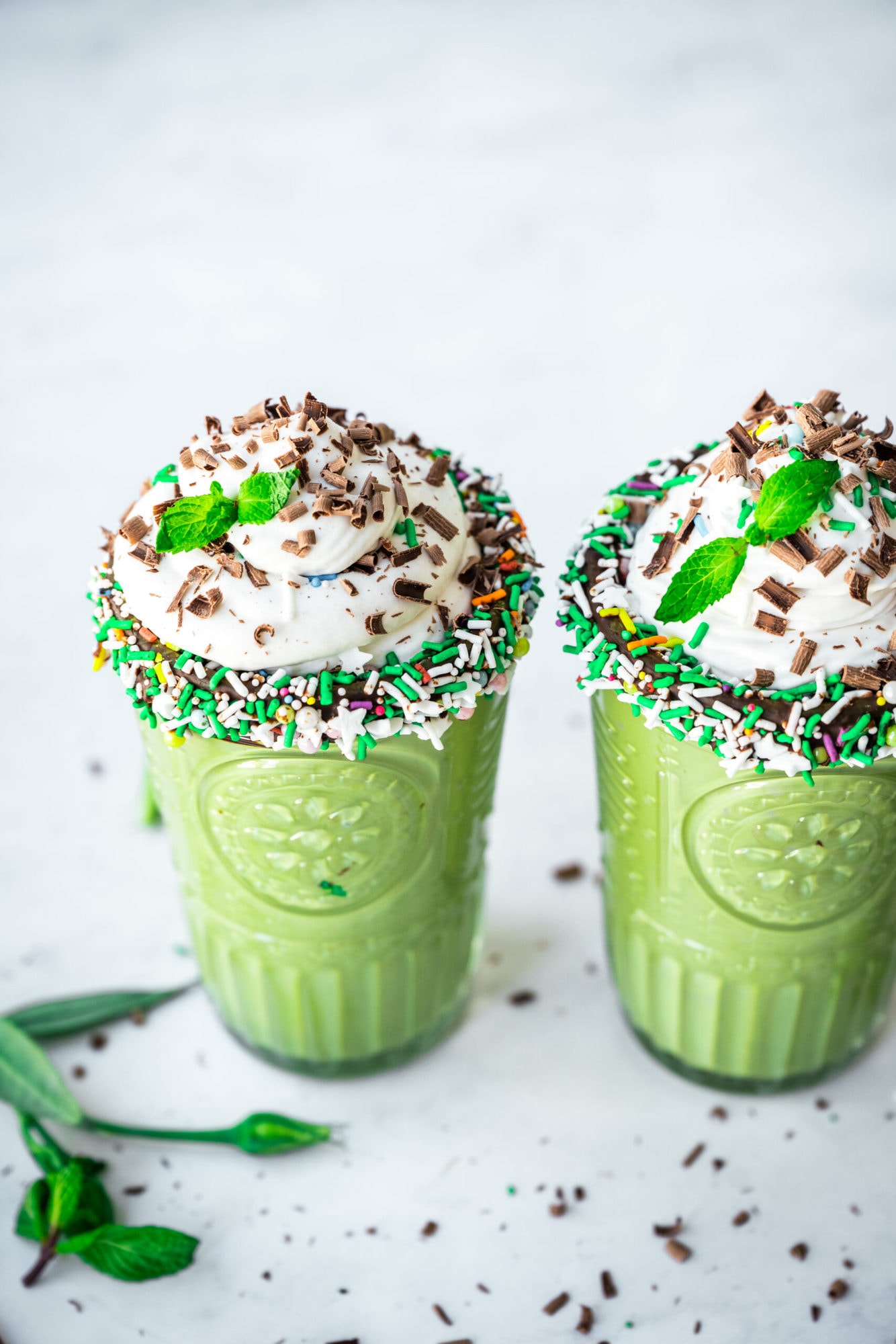 side view of two glasses of vegan shamrock shake with chocolate sprinkle rim and whipped cream on top. 